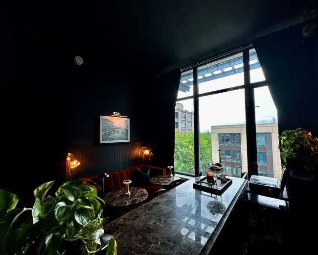 The image shows a dimly lit interior of a café or lounge with a large window overlooking greenery and neighboring buildings.