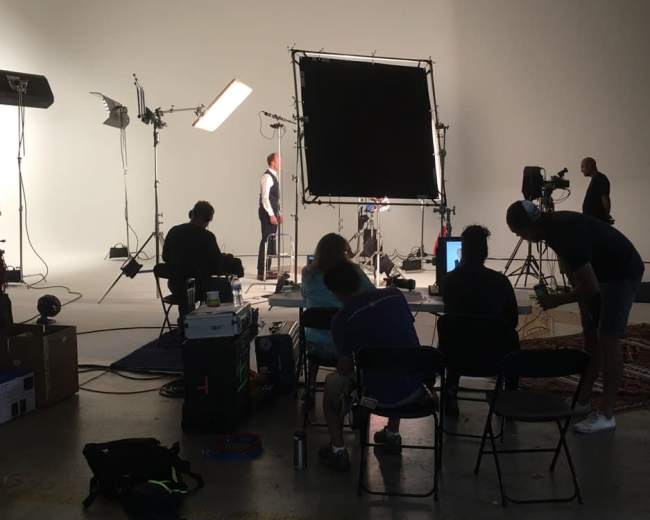 A film crew sets up equipment in a studio while people sit on chairs looking at a monitor.