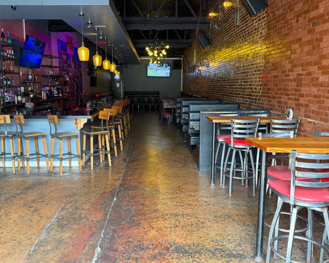 The image shows a spacious bar and restaurant with a long wooden counter, high stools, and tables arranged along a brick wall.