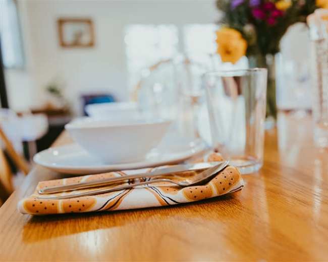 A wooden dining table is set with a folded napkin, a spoon and fork, glassware, and empty bowls, with a floral arrangement in the background.