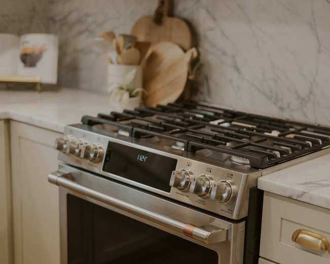 The image shows a stainless steel gas range on a marble countertop, accompanied by wooden cutting boards and kitchen utensils.