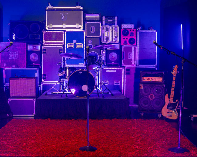 A stage setup features a drum kit surrounded by stacks of amplifiers and speakers, with three microphone stands in front and a red rug on the floor.