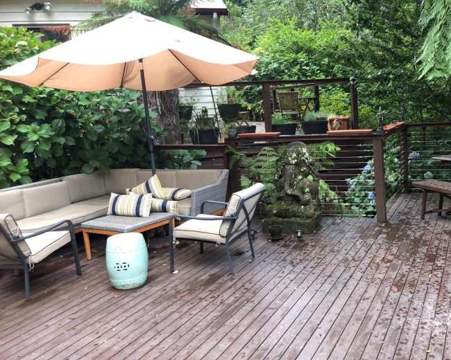 The image shows a wooden deck area with a seating arrangement under an umbrella, a small stone statue, and a dining table surrounded by lush greenery.