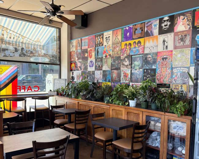 The interior of a café features wooden tables and chairs arranged near a wall decorated with various colorful album covers and houseplants.
