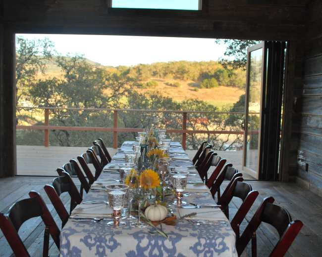 A long dining table is set with a floral centerpiece and is positioned in front of an open doorway that leads to a view of a hillside.