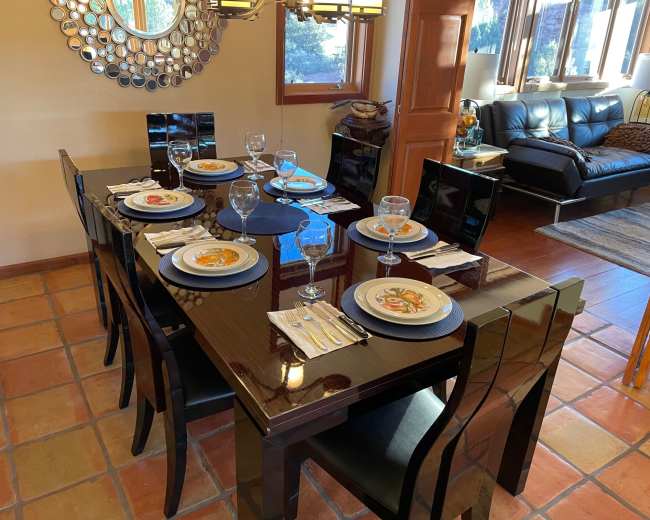 A polished black dining table is set for dinner with plates, glasses, and cutlery, surrounded by stylish chairs in a well-lit room featuring a mirrored wall and a couch in the background.