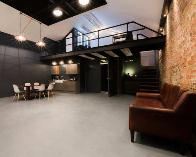 The image depicts a modern studio space featuring a lofted area, a kitchen, a dining table with chairs, a leather couch, and exposed brick walls.