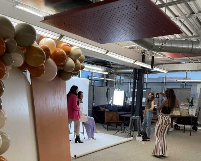Two women pose for a photo shoot in an office space decorated with balloons and a backdrop, while a photographer captures the scene.
