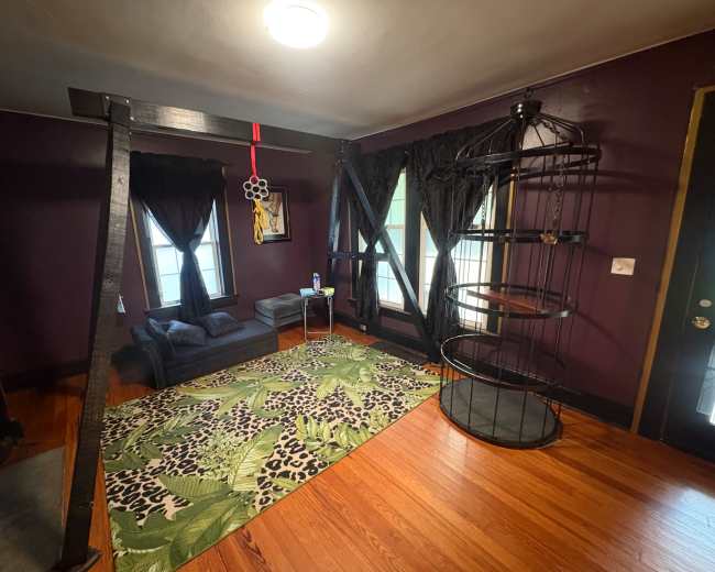 The room features dark purple walls, a patterned rug, a modern seating area with a small table, and a unique, circular shelf structure beside large windows dressed with black curtains.