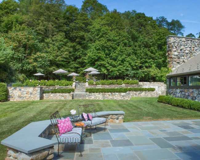 A landscaped backyard featuring a stone patio, a seating area with striped cushions, and steps leading to a garden with umbrellas and lush greenery in the background.