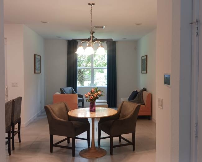 The image shows a cozy living area featuring a round dining table surrounded by four chairs, with two armchairs and a couch positioned nearby, illuminated by a hanging light fixture.