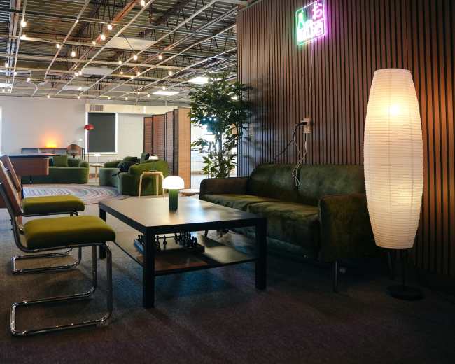 The image depicts a modern office space with green chairs, a dark coffee table, a sofa, and a floor lamp, surrounded by plants and a neon sign.