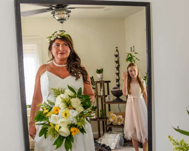 A bride in a white dress stands in front of a mirror holding a bouquet of flowers, while a young girl in a light dress appears in the background.