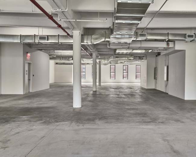 An empty commercial space with exposed ductwork, concrete floors, and several windows along the walls.