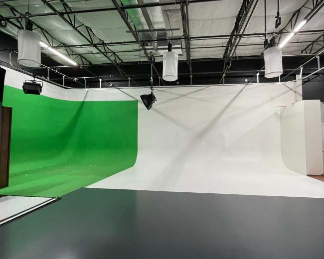 A partially empty studio space with a green screen on one side and a white background on the other, under a ceiling with hanging lights.