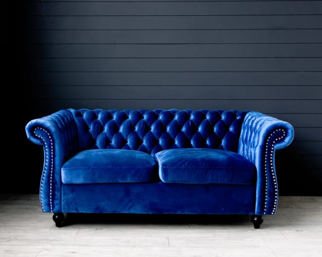 A blue velvet chesterfield sofa with tufted upholstery and wooden legs is positioned against a dark wall.