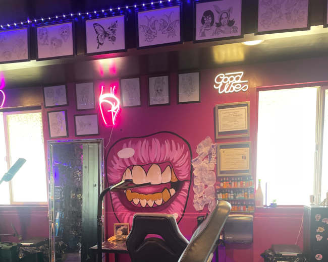 The image shows a tattoo parlor interior featuring a tattoo chair, vibrant pink walls adorned with artwork, and a neon sign that reads "Good Vibes."