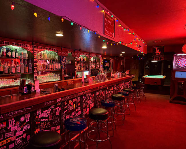 The image shows a dimly lit bar with a long counter, illuminated by colorful string lights, featuring shelves stocked with bottles and a pool table in the corner.