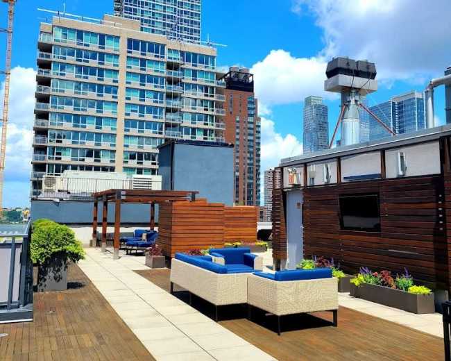 The image shows a rooftop terrace with wooden decking, lounge seating, and city skyscrapers in the background under a blue sky with scattered clouds.