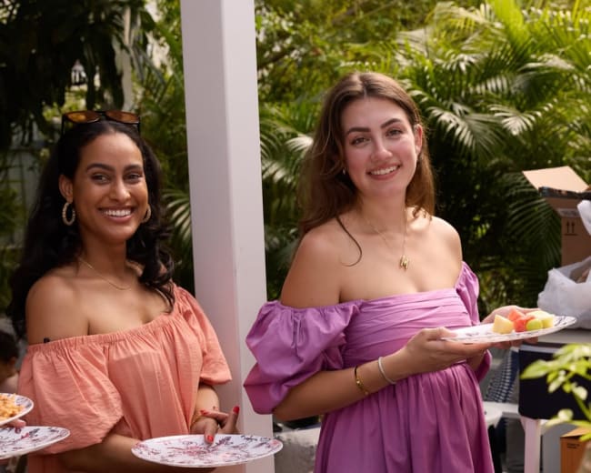 Two women wearing colorful, off-the-shoulder dresses hold plates of food while standing in a garden setting.