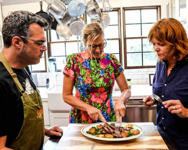 Three people are in a kitchen, with one woman slicing meat on a plate while the others watch attentively.