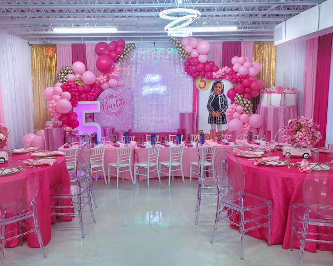 A brightly decorated birthday party scene with pink and white balloons, tables covered in pink tablecloths, and a large "5" centerpiece surrounded by themed decorations.