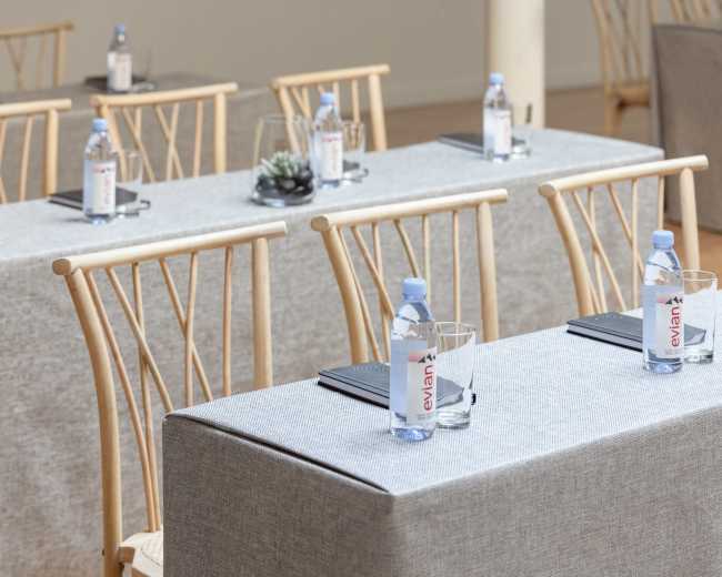 A conference setup features several tables covered in gray linen, each adorned with a bottle of Evian water, a glass, and a small plant.