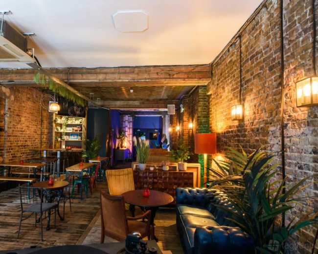 The image shows the interior of a cozy café with exposed brick walls, wooden beams, and a mix of seating arrangements, including tables and a leather couch.