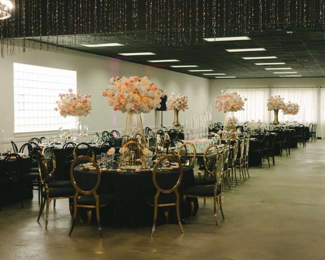 A spacious banquet hall features round tables adorned with black tablecloths and tall floral arrangements, set for a formal event.