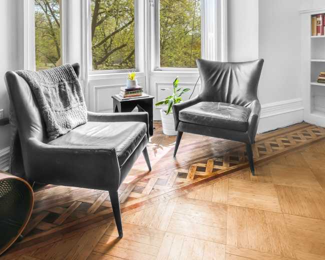 The image shows a bright corner of a room with two black armchairs positioned near large windows overlooking trees outside.