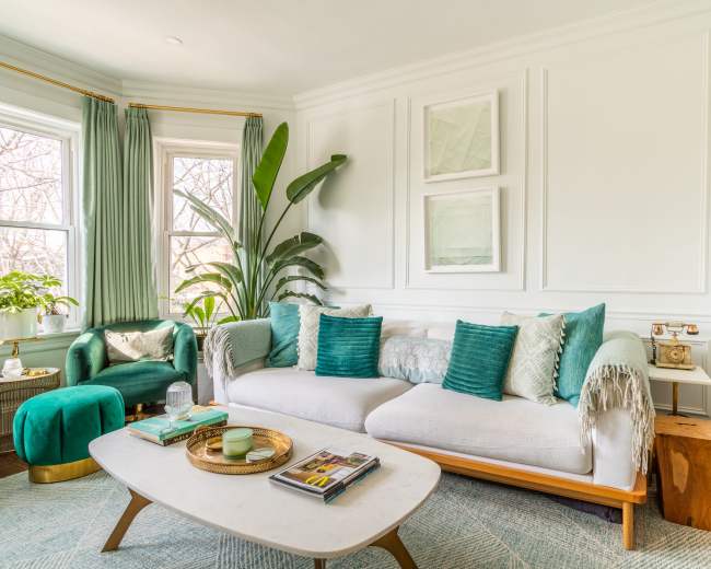A living room features a light gray sofa with teal cushions, a round green ottoman, large windows with green curtains, and various houseplants.