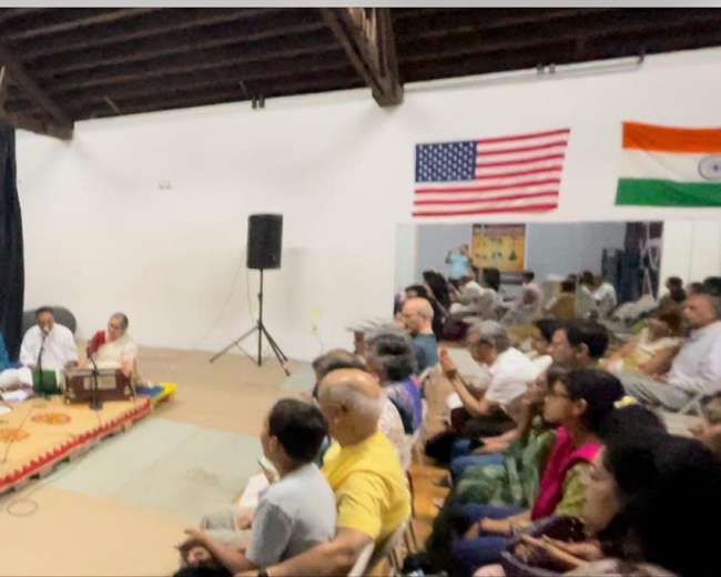 A group of musicians performs on a stage in front of an audience in a decorated hall featuring Indian and American flags.