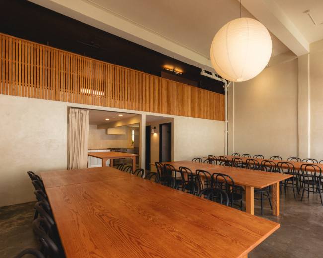 The image shows a spacious dining area with long wooden tables and black chairs, illuminated by a large pendant light.