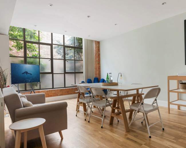 The room features a modern dining area with a wooden table, folding chairs, and a large window displaying a greenery view.