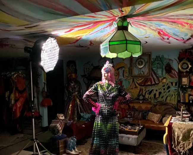 A person in a colorful, patterned outfit stands in a room with a checkered floor, surrounded by various costumes and decorated walls.
