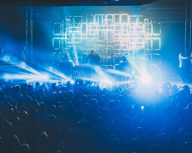 A large crowd gathers in a dimly lit venue as a performer plays electronic music behind a backdrop of illuminated geometric lights.