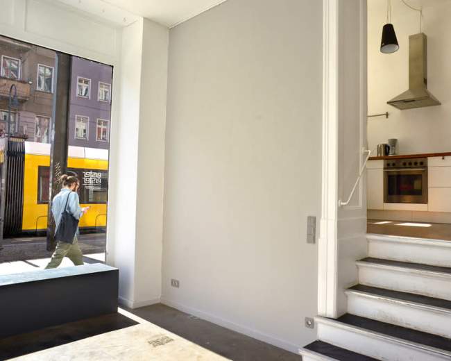 A person walks by a large window in a minimalist interior featuring an open staircase and a kitchen area.