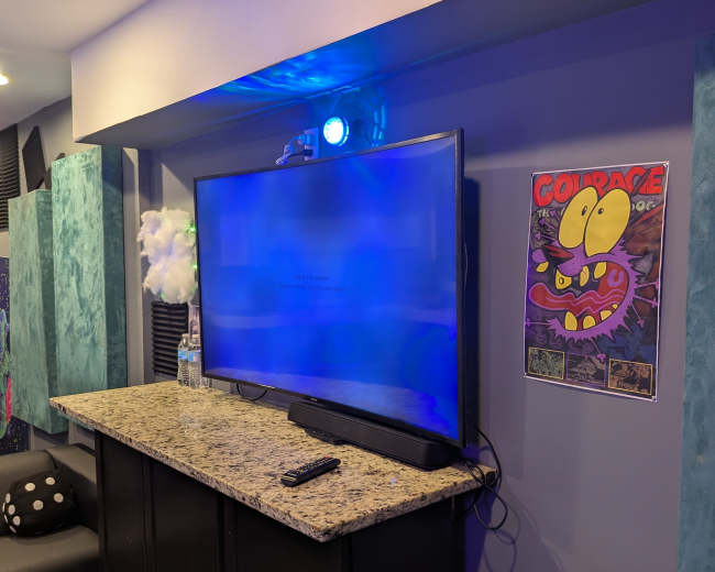 The image shows a modern entertainment setup featuring a large flat-screen TV mounted on a wall above a granite countertop, with a colorful poster and a remote control placed on the surface below.