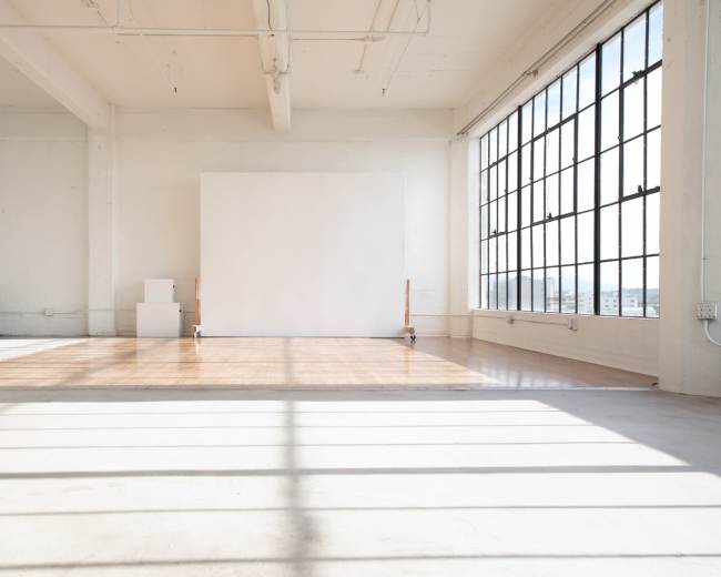 A spacious, bright room with large windows, featuring a polished wooden floor and a blank white wall at one end.