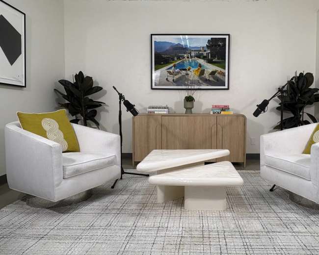 The image shows a contemporary living space with two white armchairs, a sculptural coffee table, a wooden sideboard, and decorative plants, all arranged on a patterned rug.