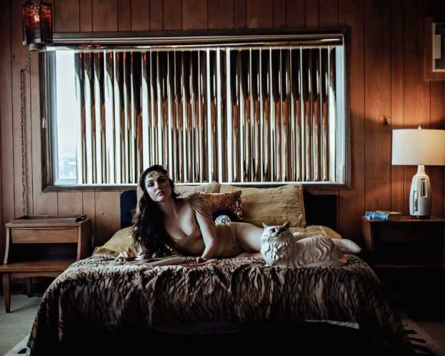 A woman lies on a bed with a tiger-striped cover, next to a white cat, in a room featuring wood-paneled walls and a window with vertical blinds.