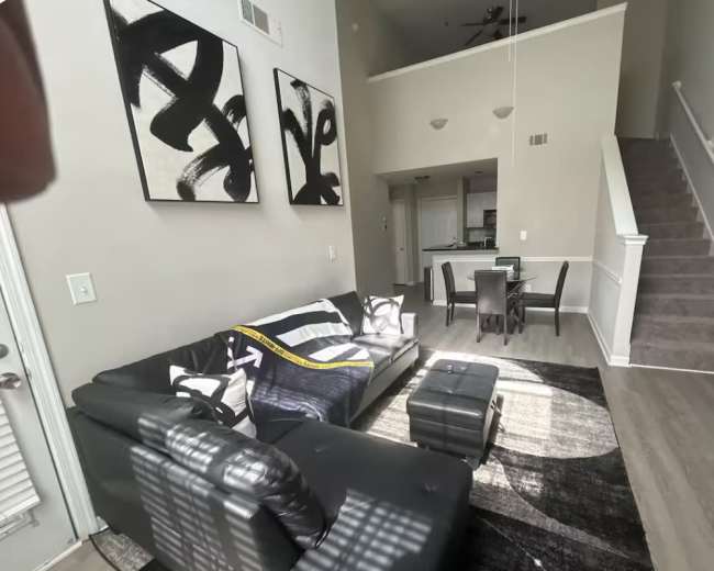 The image shows a modern living room with a black sectional sofa, two abstract wall art pieces, and a staircase leading to an upper level.