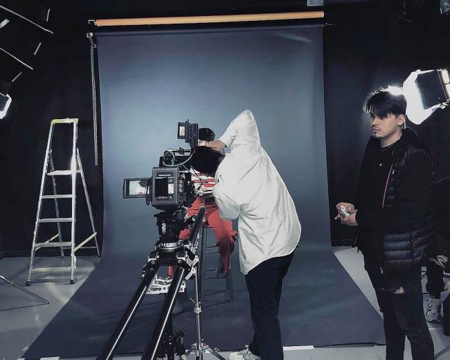 A person in a white jacket operates a camera on a tracking dolly while another individual stands nearby in a dimly lit studio with a backdrop and lighting equipment.