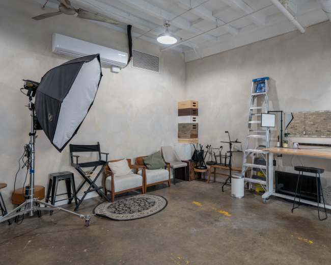 The image shows a photography studio with a backdrop, seating area, a ladder, and equipment storage on the walls.