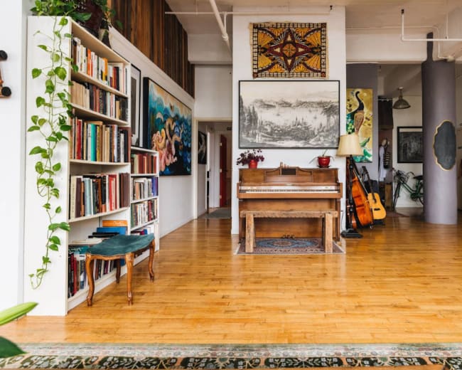 The image shows a spacious room with hardwood floors, featuring a piano, bookshelves filled with books, and various artistic decorations on the walls.