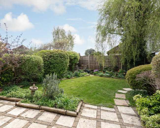 A well-maintained garden features a grassy area surrounded by neatly trimmed hedges and flower beds, with a stone walkway leading through the space.