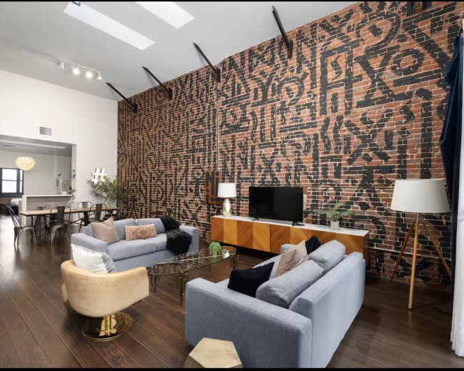 The image shows a modern living room with a brick wall featuring black abstract patterns, two sofas, a coffee table, and a television on a wooden media console.