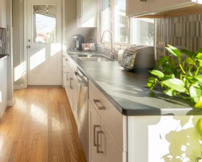 The kitchen features white cabinetry, a countertop, and a door leading outside, illuminated by natural light.