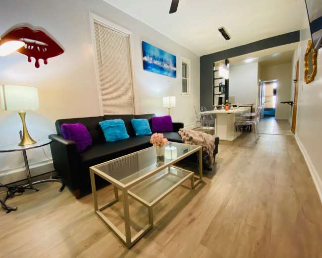A modern living room with a black sofa, colorful cushions, a glass coffee table, and a dining area visible in the background.