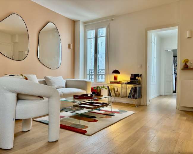 A modern living room features a light-colored sofa, unique-shaped mirrors, a glass coffee table, and a blend of minimalistic decor and natural light.